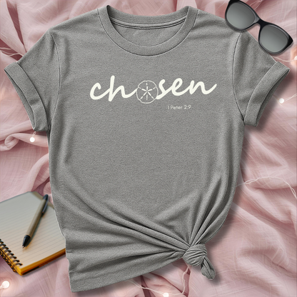 Gray t-shirt with 'chosen' text on a pink surface with sunglasses and notebook.