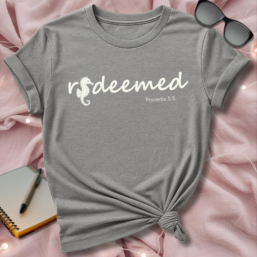 Gray t-shirt with 'redeemed Proverbs 3:5' text on a pink surface with sunglasses and notebook.