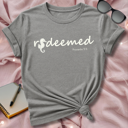Gray t-shirt with 'redeemed Proverbs 3:5' text on a pink surface with sunglasses and notebook.