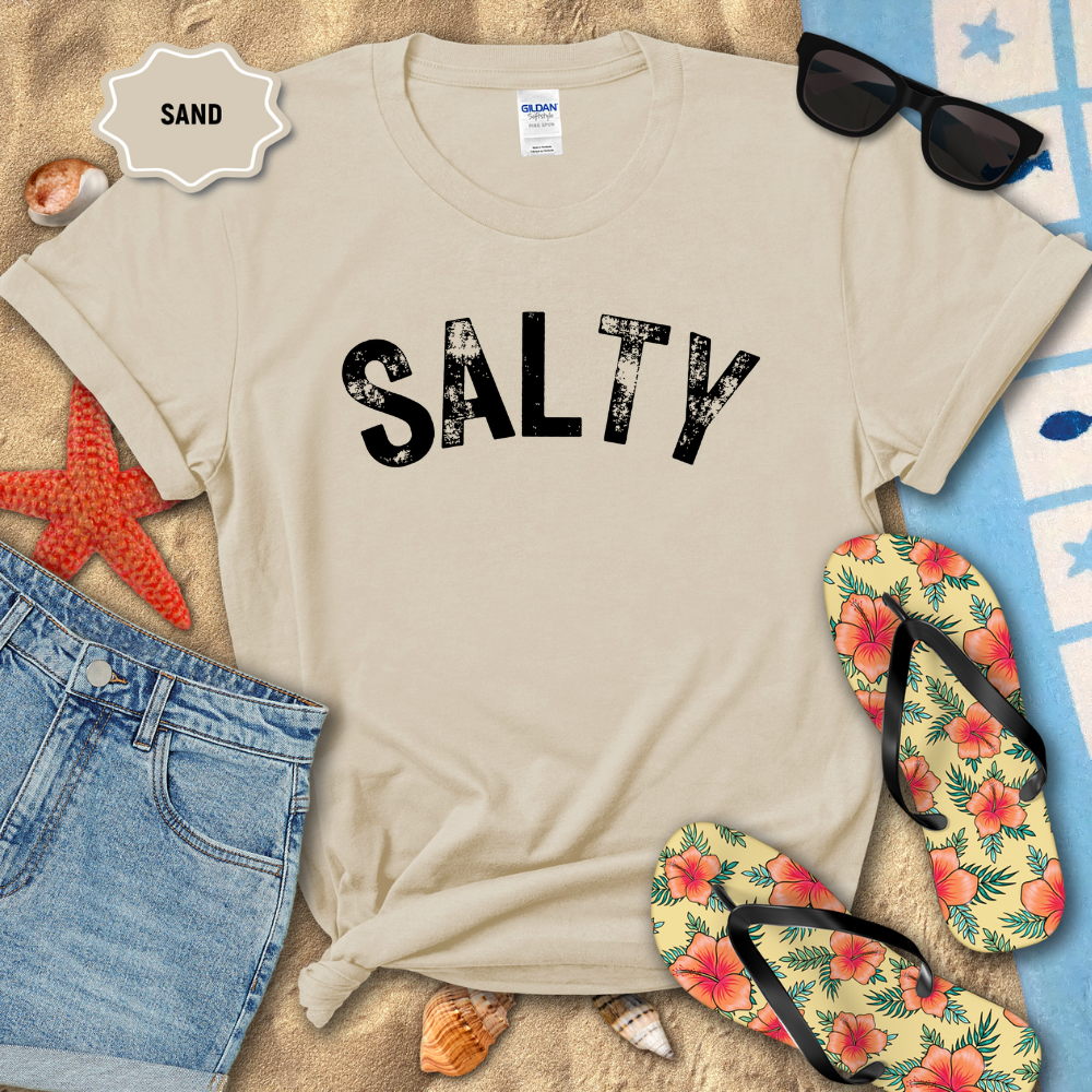 Beige t-shirt with 'SALTY' text on a sandy background with flip-flops and sunglasses.