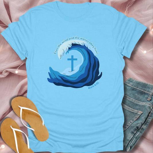 Light blue t-shirt with wave design and cross, placed on a pink fabric background with sandals and jeans.