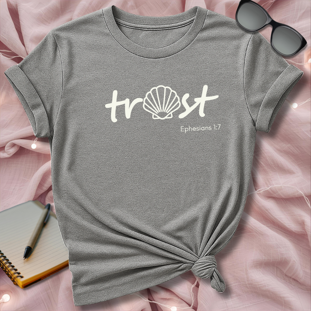 Gray t-shirt with 'trust' and shell design on a pink surface with sunglasses and notebook.