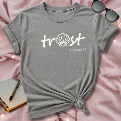 Gray t-shirt with 'trust' and shell design on a pink surface with sunglasses and notebook.