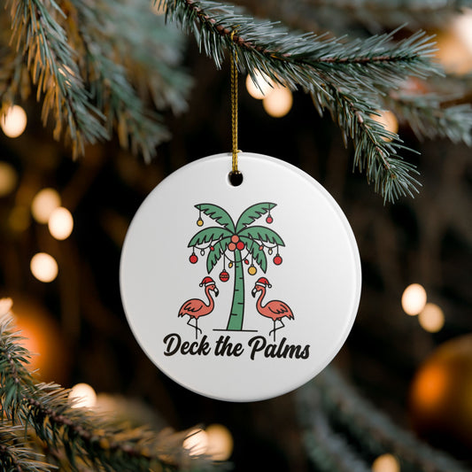 A round white Christmas ornament hanging from a pine tree branch, featuring a green palm tree with red ornaments, two pink flamingos, and the text 'Deck the Palms' against a blurred holiday tree background with warm lights.