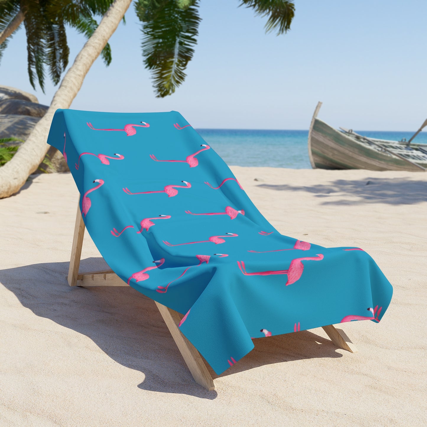 Blue beach towel with pink flamingo pattern draped over a lounge chair on a sandy beach.