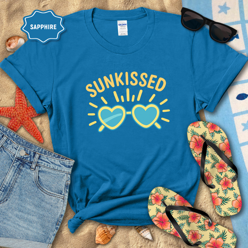 Blue t-shirt with 'SUNKISSED' design on a beach setting with sunglasses, flip-flops, and denim shorts.