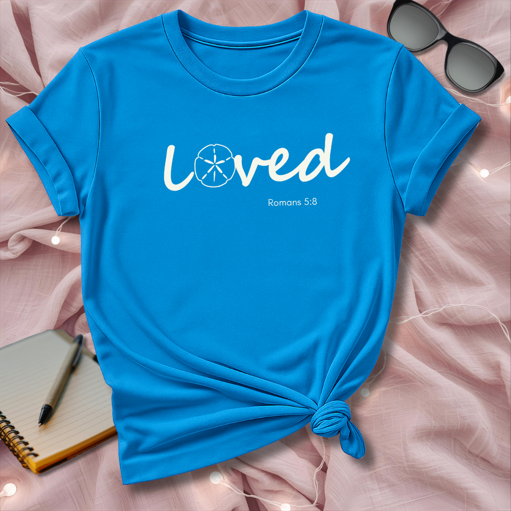 Blue t-shirt with 'loved' text on a pink surface with sunglasses and notebook.