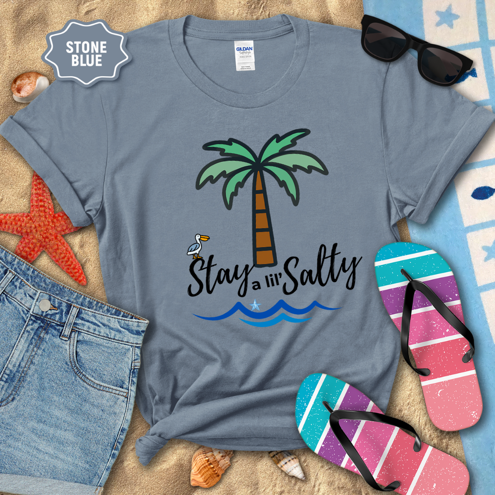 Stone blue t-shirt with palm tree and 'Stay a lil' Salty' text, surrounded by beach accessories.