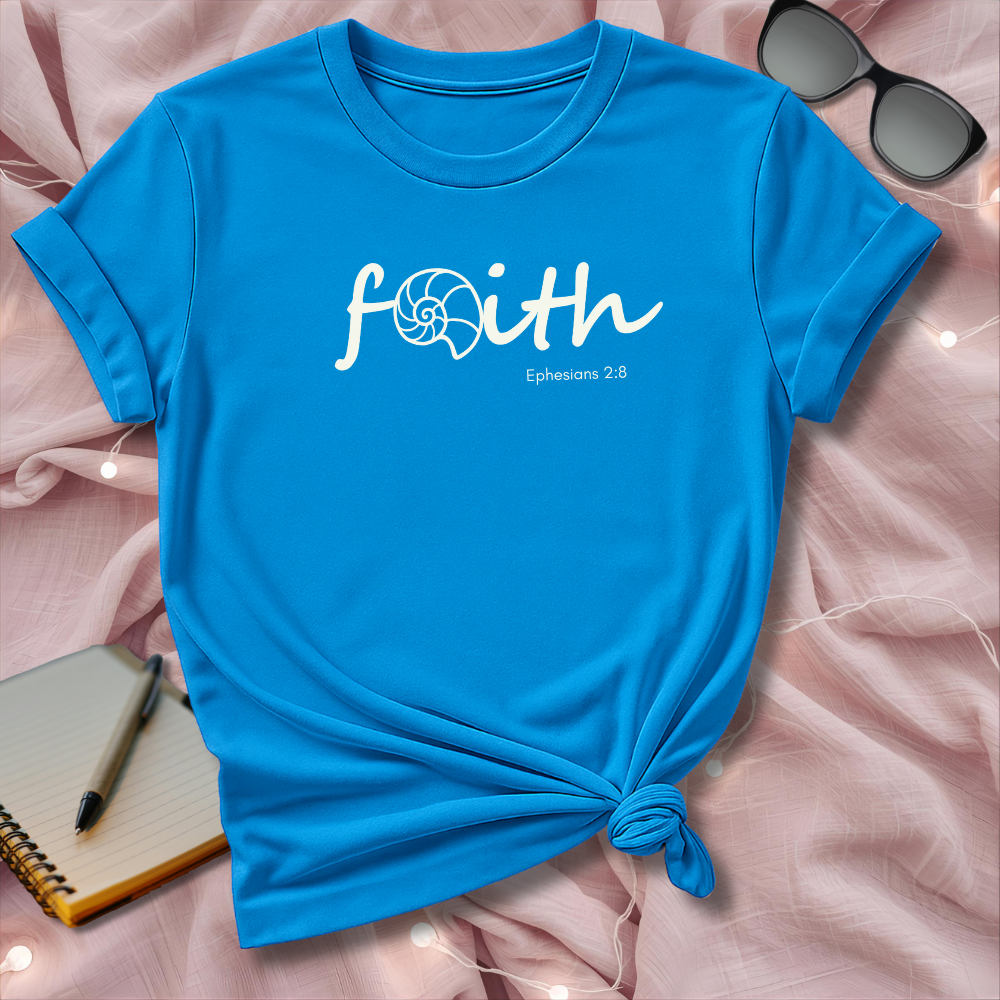 Blue t-shirt with 'faith' text on a pink surface with sunglasses and notebook.