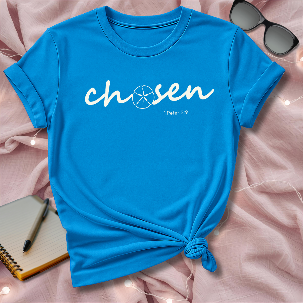 Blue t-shirt with 'chosen' text on a pink surface with sunglasses and notebook.