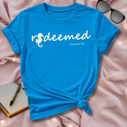 Blue t-shirt with 'redeemed' text on a pink surface with sunglasses and notebook.