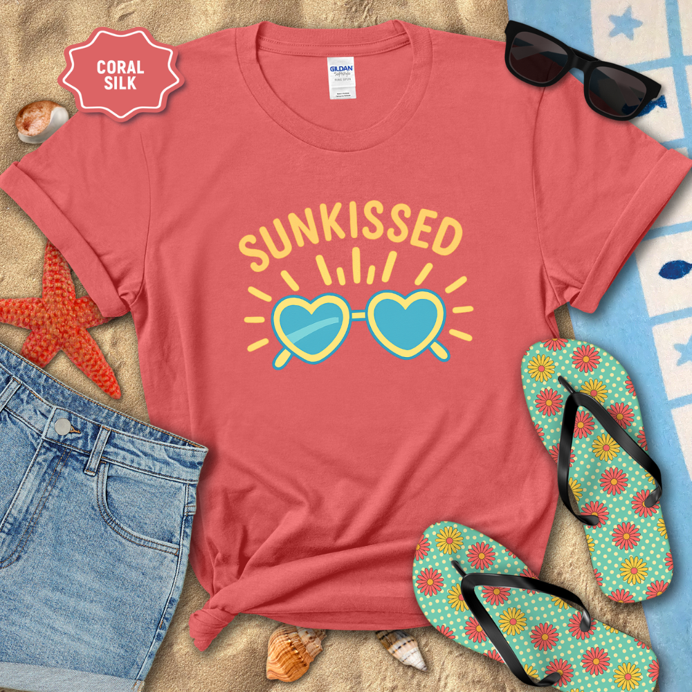 Coral t-shirt with 'Sunkissed' design on a sandy surface with sunglasses and flip-flops.