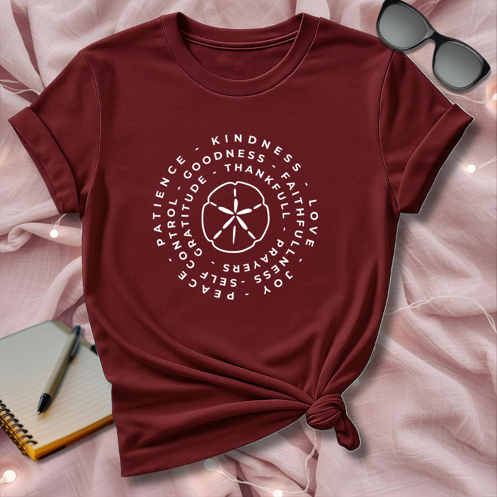 Maroon t-shirt with white text and design on a pink surface with sunglasses and notebook.