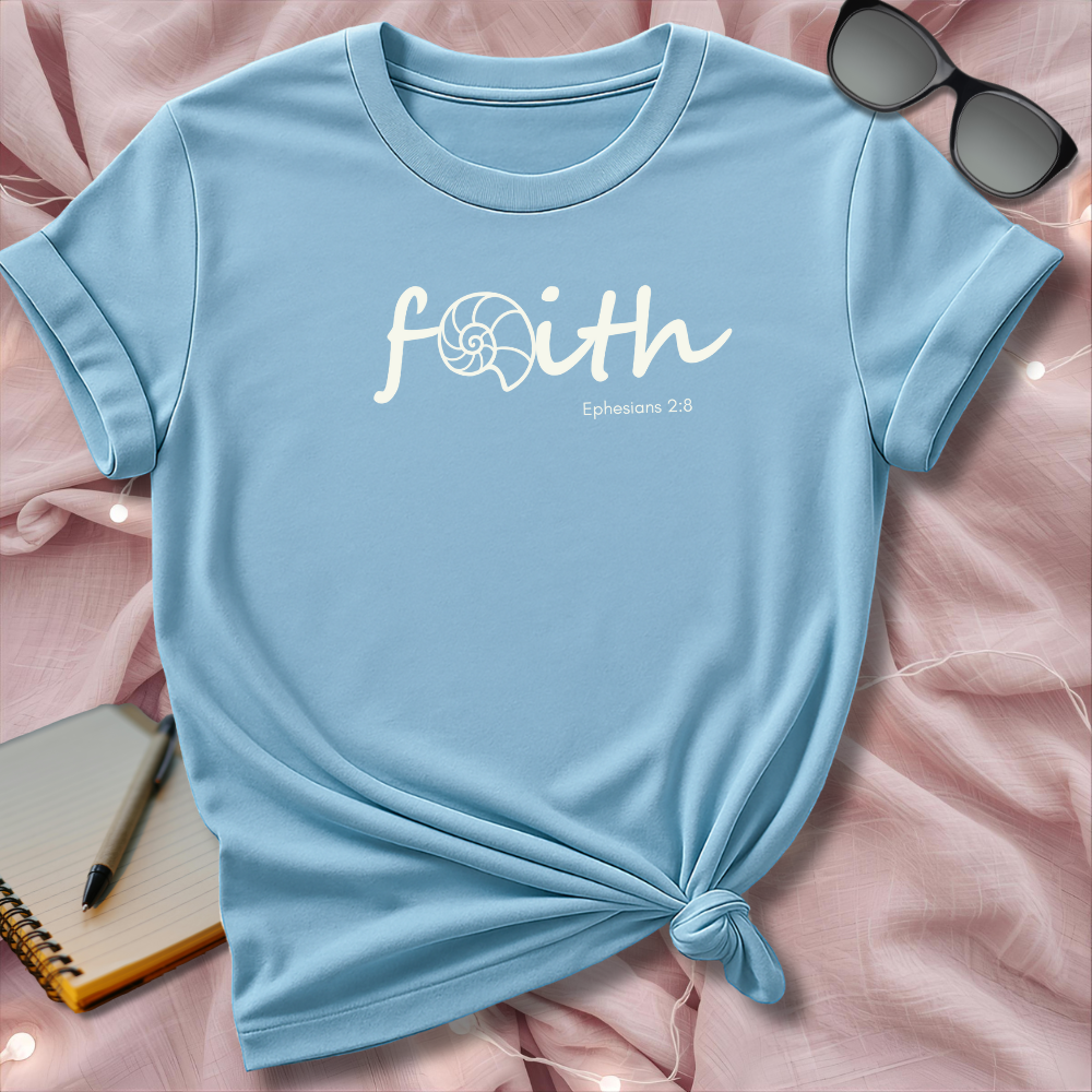 Light blue t-shirt with 'faith' text on a pink surface with sunglasses and notebook.