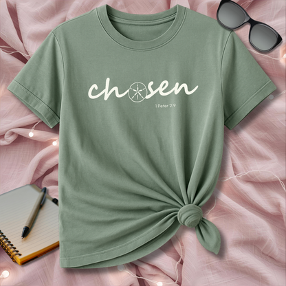 Green t-shirt with 'chosen' text on a pink surface with sunglasses and notebook.