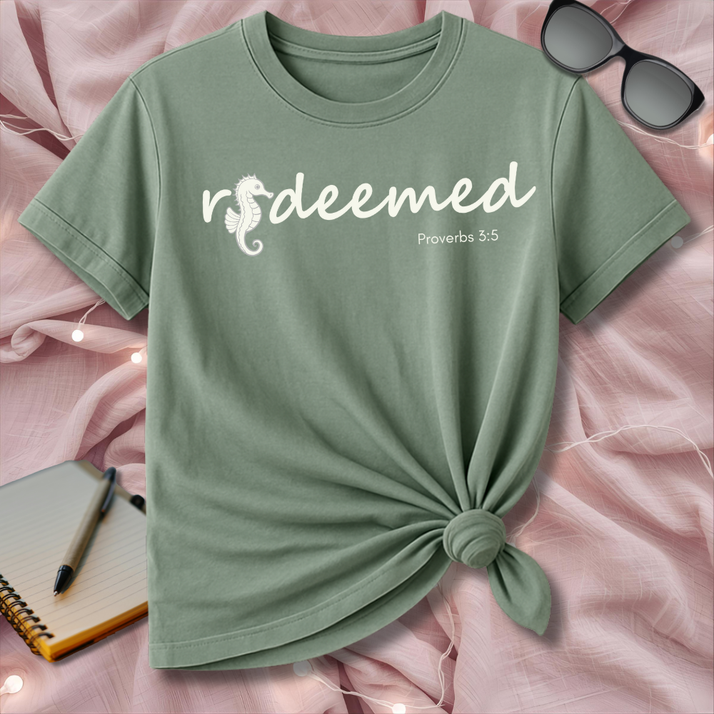 Green t-shirt with 'redeemed' text on a pink surface with sunglasses and notebook.