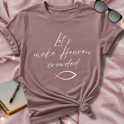 Pink t-shirt with 'Let's make Heaven crowded' text and fish symbol on a pink surface with sunglasses and notebook.