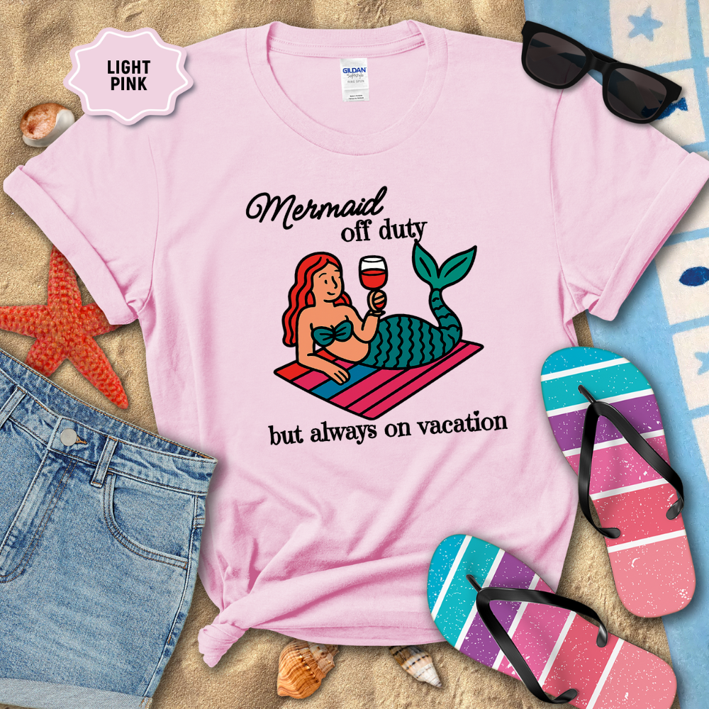 Light pink t-shirt with mermaid graphic and text, surrounded by beach accessories on sand.