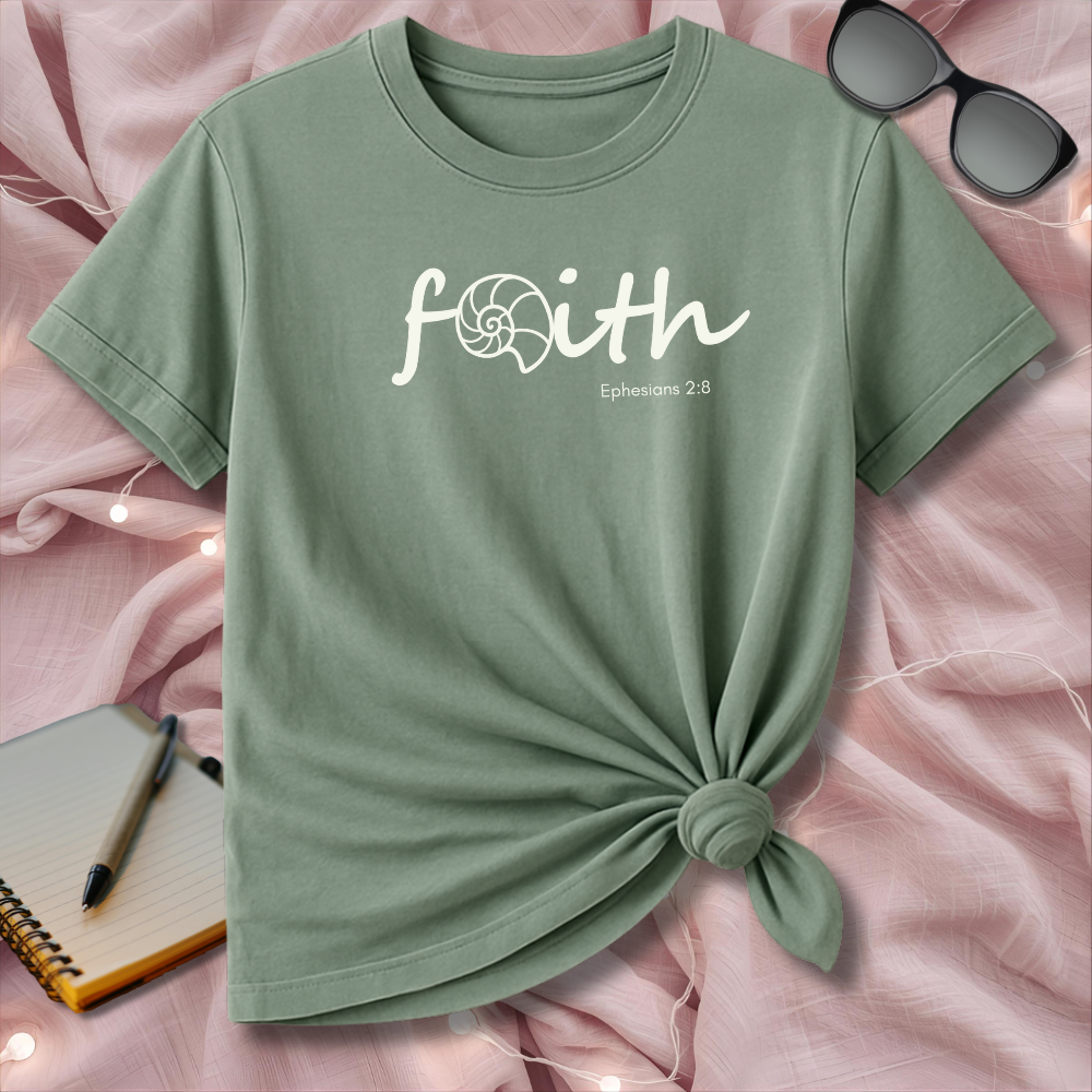Green t-shirt with 'faith' text on a pink surface with sunglasses and notebook.
