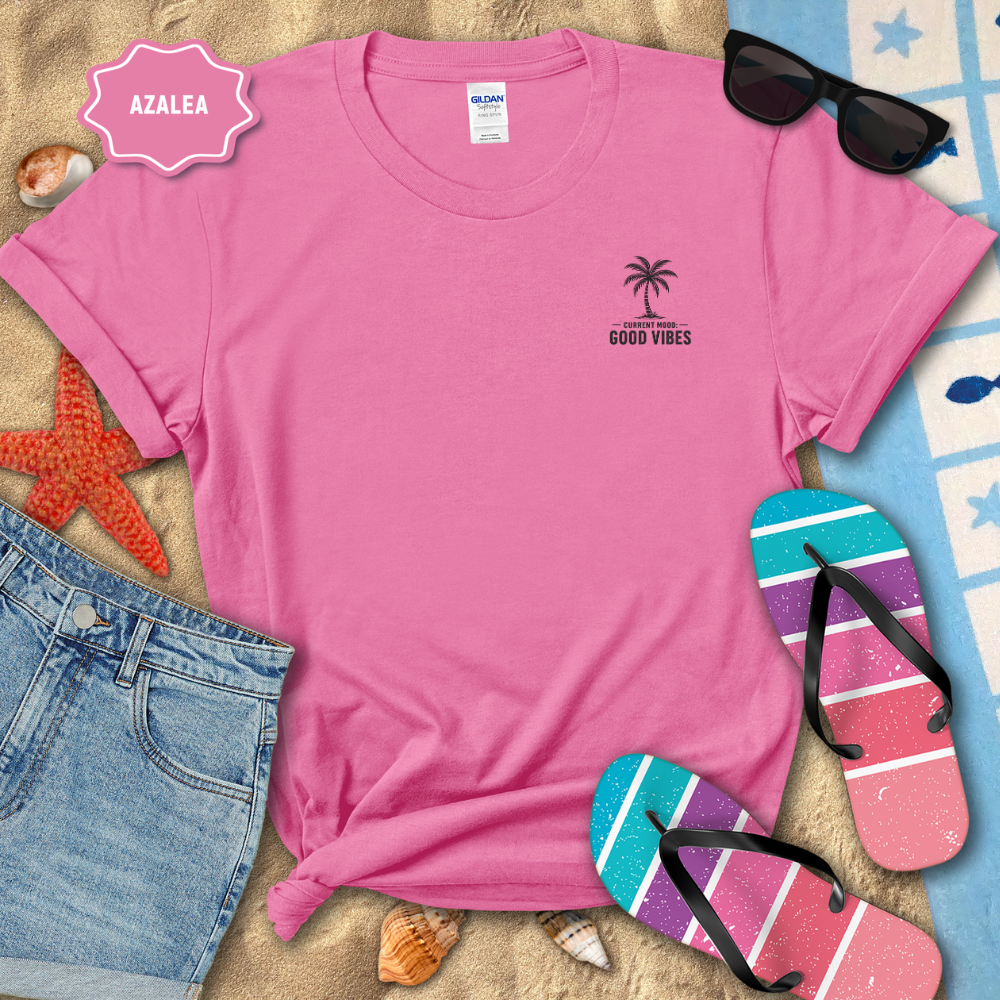 Pink t-shirt with palm tree design and 'Good Vibes' text, surrounded by beach accessories on sand.