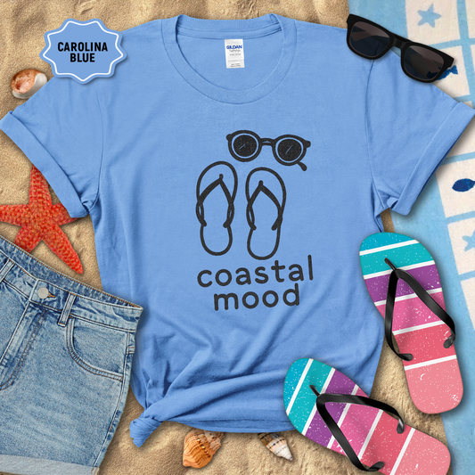 Blue t-shirt with 'coastal mood' design, sunglasses, flip-flops, and beach accessories on sand.
