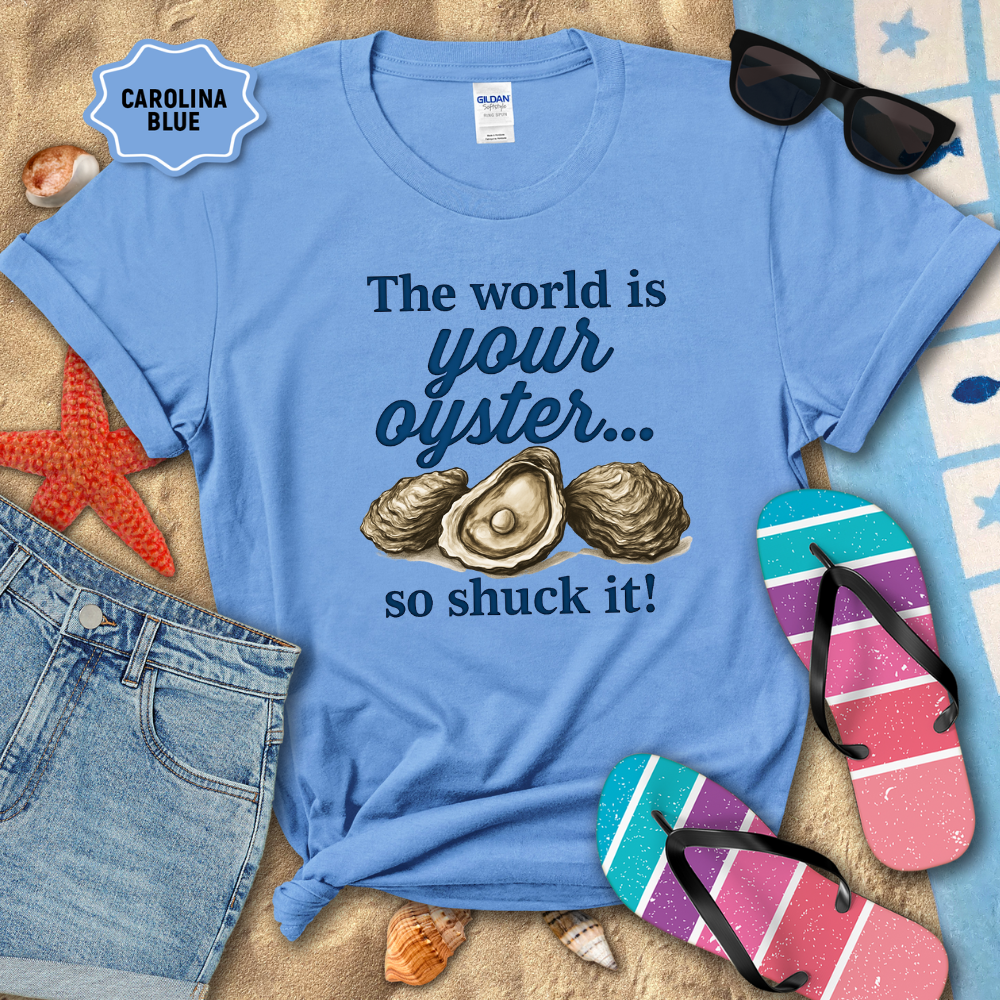 Blue t-shirt with oyster graphic and text on a sandy background with beach accessories.