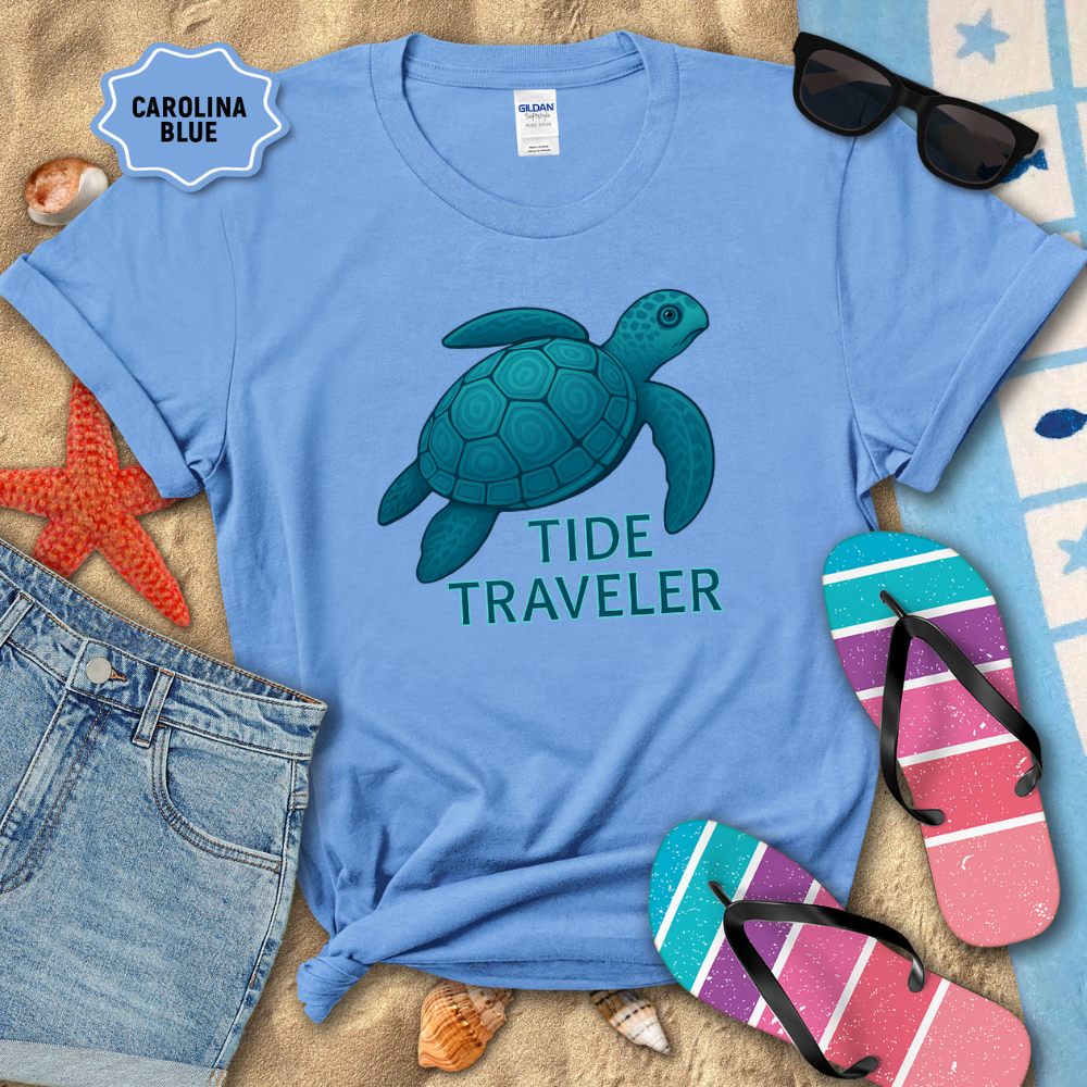 Blue t-shirt with a turtle graphic and 'Tide Traveler' text, surrounded by beach accessories on sand.