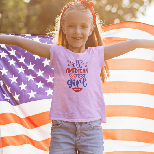 Smiling young girl wearing a pink "All American Girl" T-shirt in front of a large American flag, celebrating Independence Day.