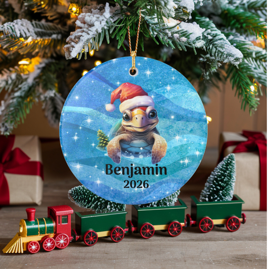 A personalized round Christmas ornament hanging from a tree branch, featuring a cartoon turtle wearing a Santa hat on a blue starry background with the name 'Benjamin 2026'. In front of the ornament is a small red, green, and gold toy train, with wrapped gifts and a Christmas tree in the background.