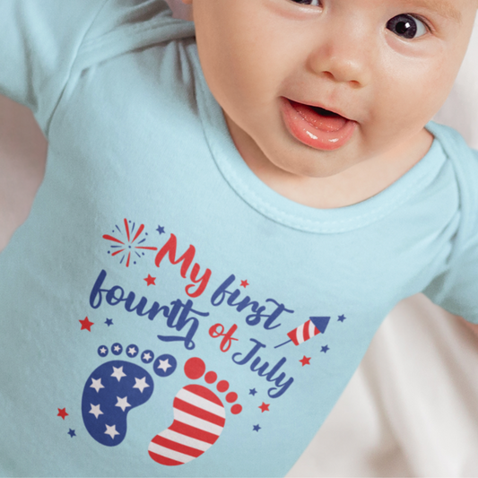 Smiling baby wearing a light blue bodysuit with “My First Fourth of July” design featuring patriotic baby footprints and fireworks.