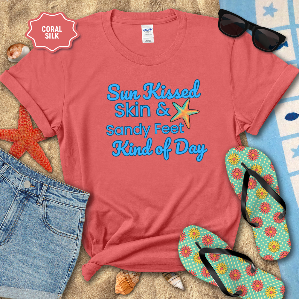 Coral t-shirt with 'Sun Kissed Skin & Sandy Feet Kind of Day' text, sunglasses, flip-flops, and beach items on sand.