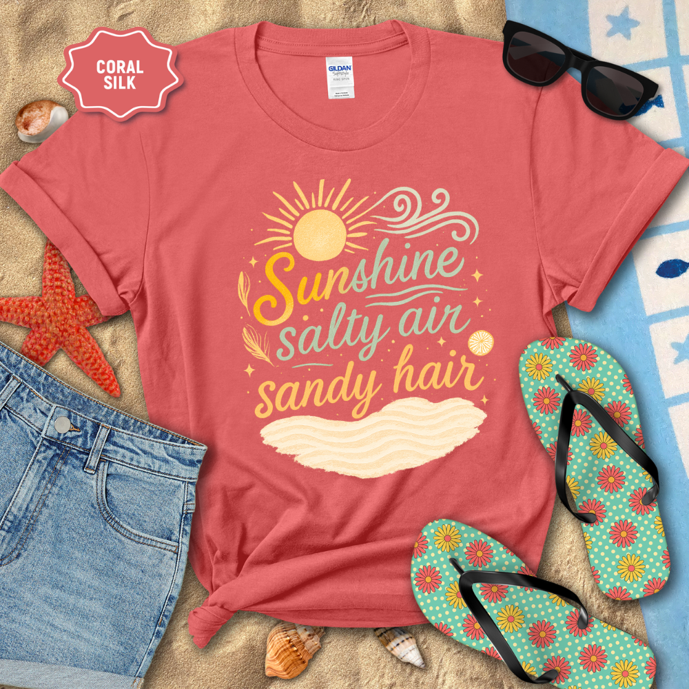 Coral t-shirt with 'Sunshine salty air sandy hair' text, sunglasses, flip-flops, and beach accessories on a sandy background.