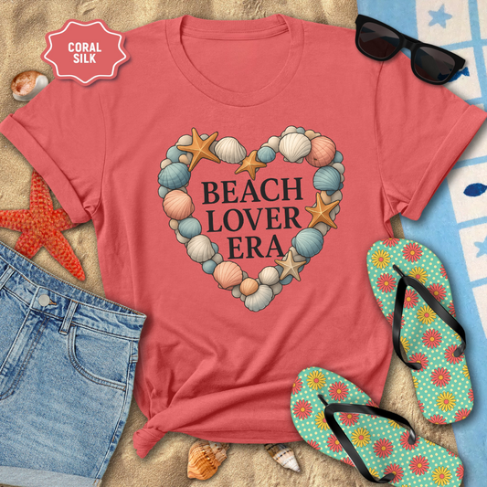 Coral t-shirt with 'Beach Lover Era' design on a sandy background with sunglasses, flip-flops, and beach accessories.