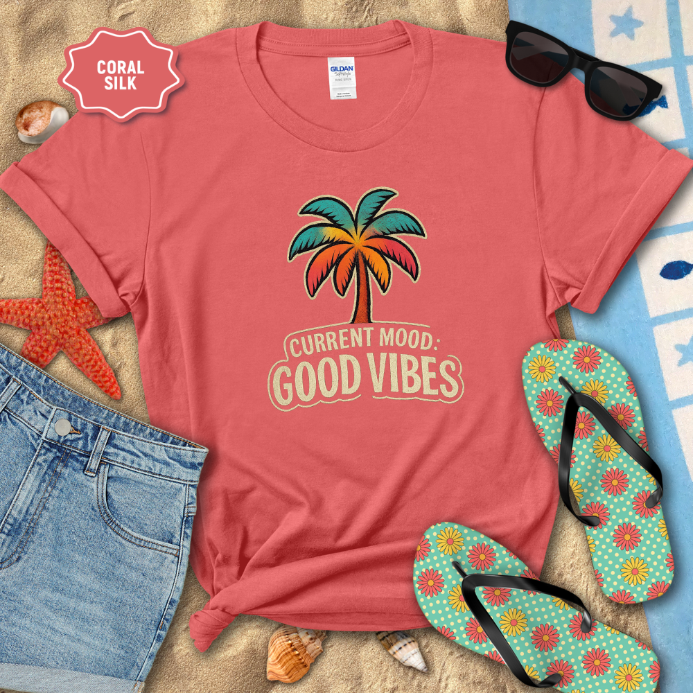 Coral t-shirt with palm tree design and 'Current Mood: Good Vibes' text, surrounded by beach accessories.