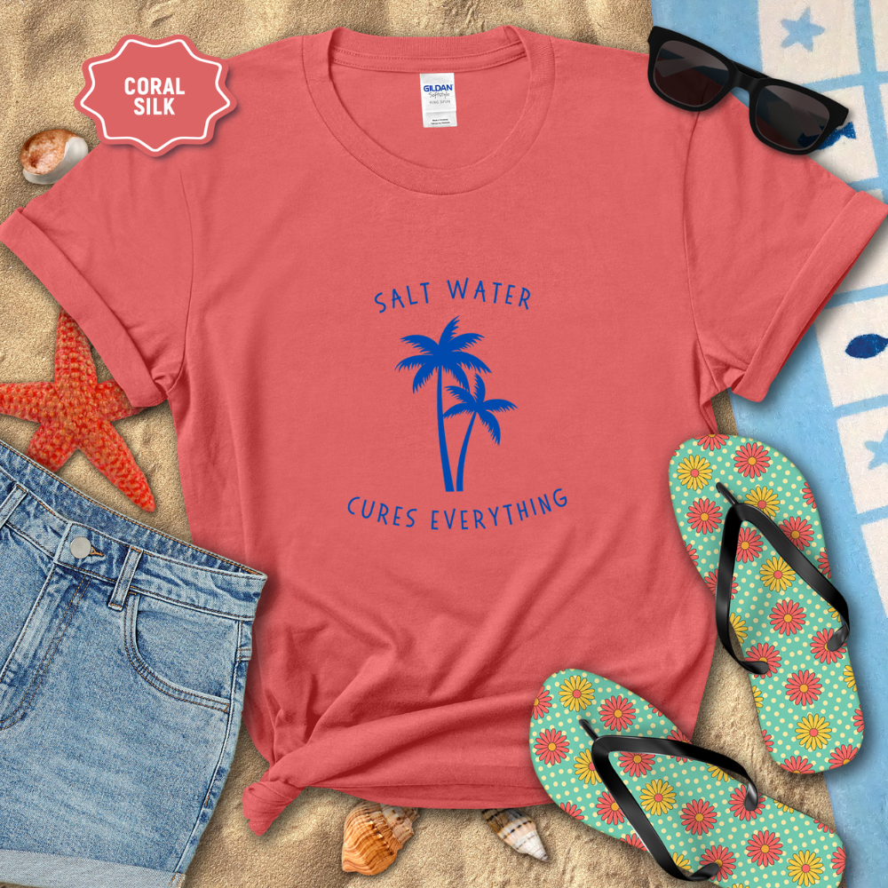 Coral t-shirt with palm tree design and text, surrounded by beach accessories on sand.