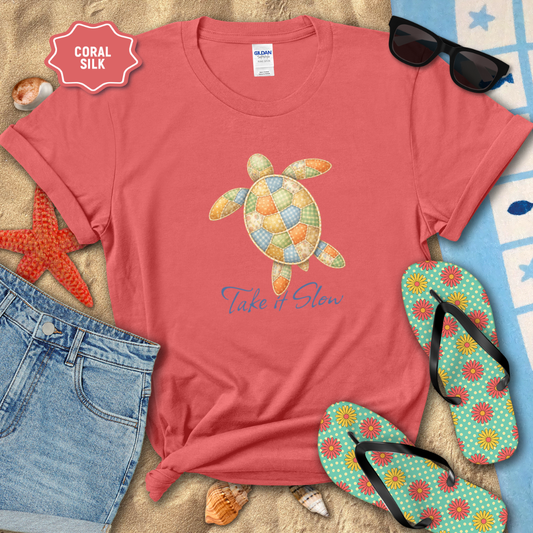Red t-shirt with a turtle design and 'Take It Slow' text, surrounded by beach accessories on sand.
