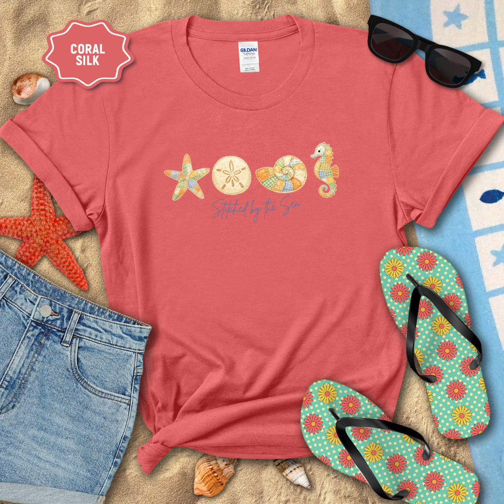Coral t-shirt with beach-themed design on sand with sunglasses, flip-flops, and starfish.