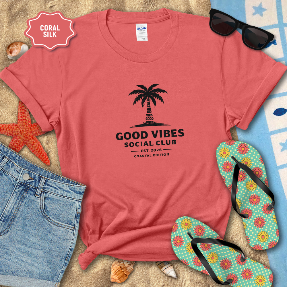 Coral t-shirt with 'Good Vibes Social Club' design on a beach setting with sunglasses, flip-flops, and beach accessories.