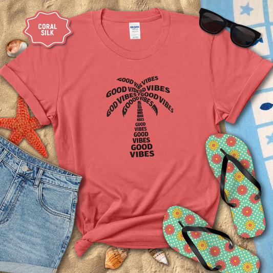 Coral t-shirt with 'Good Vibes' text, sunglasses, flip-flops, and beach items on sand.