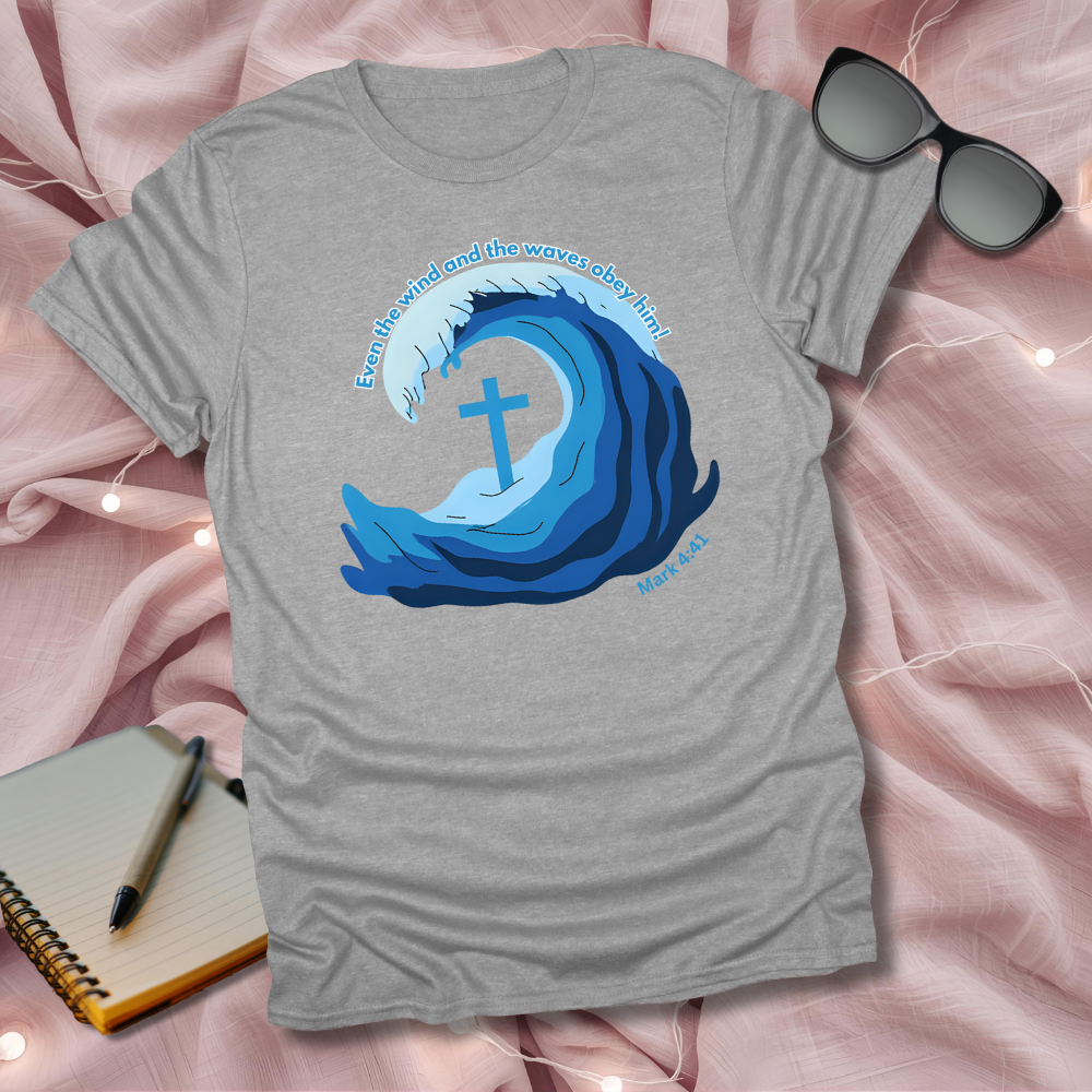 Gray t-shirt with blue wave design and cross, placed on a pink fabric background with sunglasses and notebook.