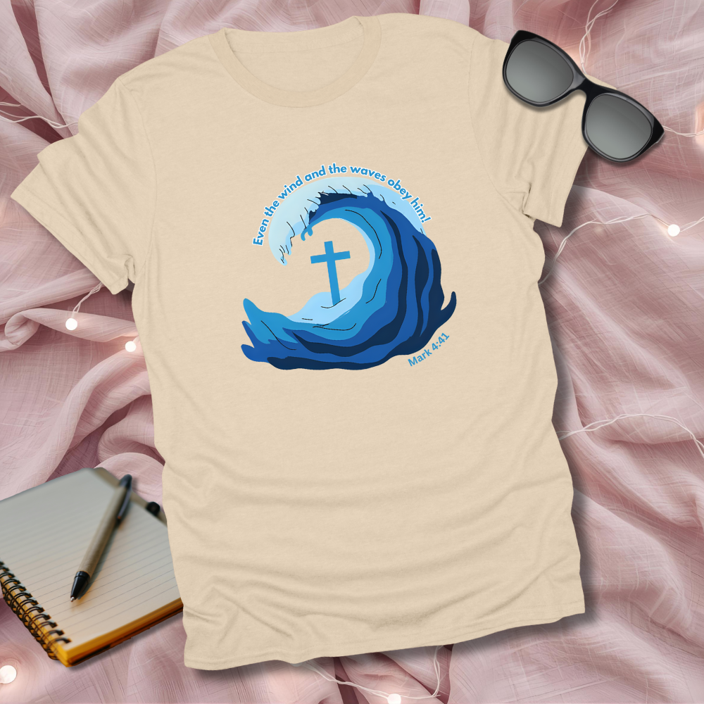 Beige t-shirt with blue wave design and cross, placed on a pink surface with sunglasses and notebook.