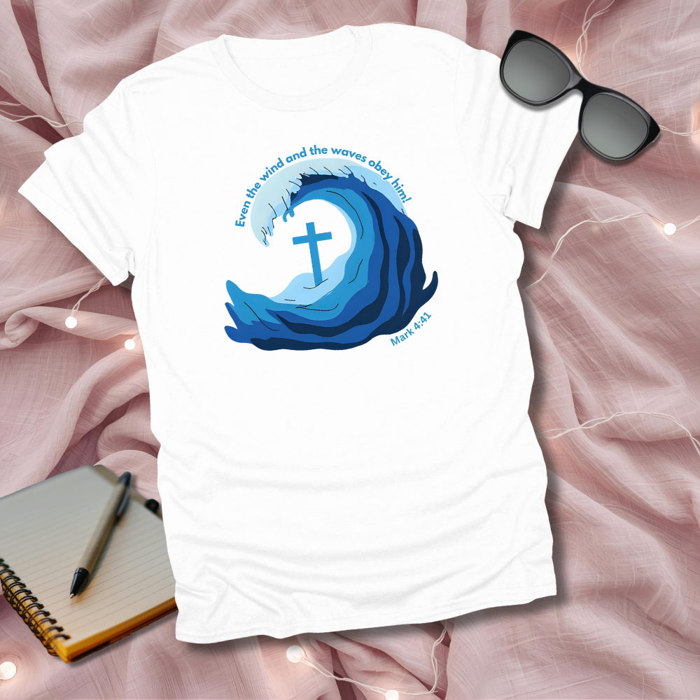 White t-shirt with blue wave design and cross, placed on a pink fabric background with sunglasses and notebook.