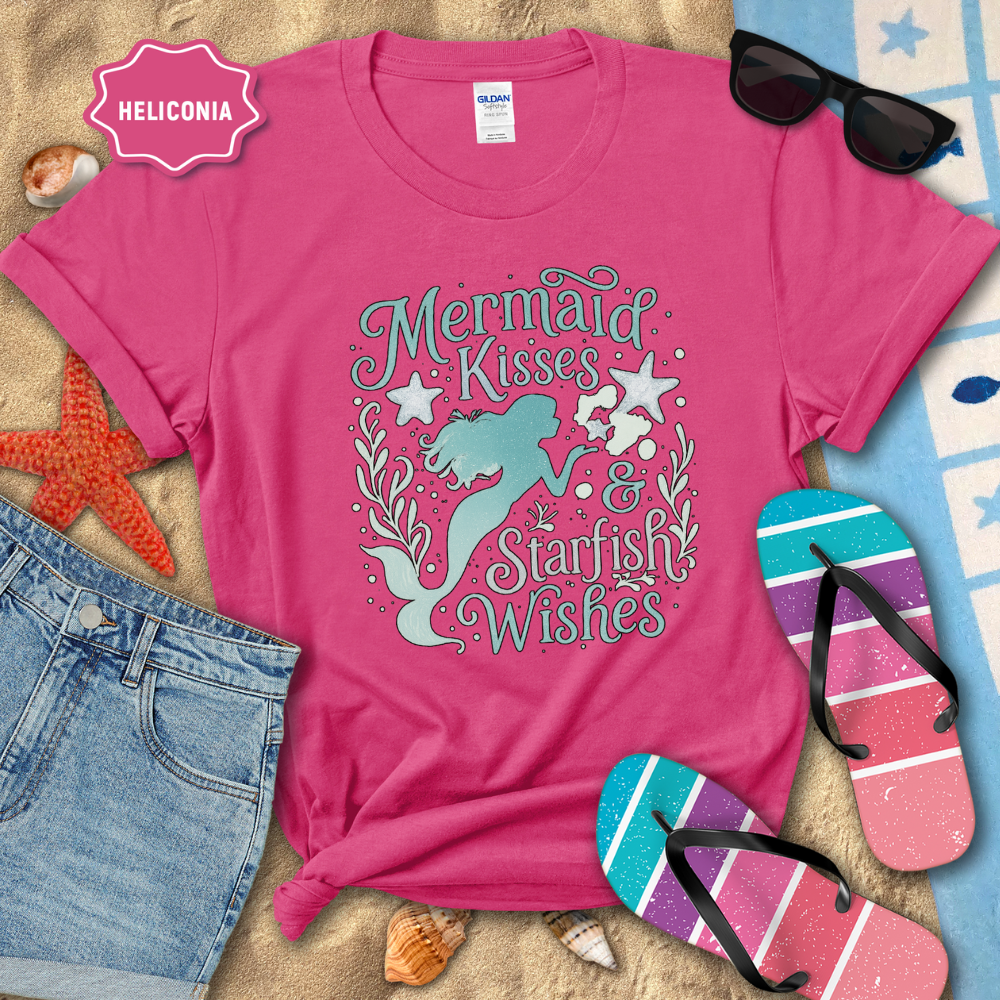 Pink t-shirt with mermaid design and text, surrounded by beach accessories on sand.