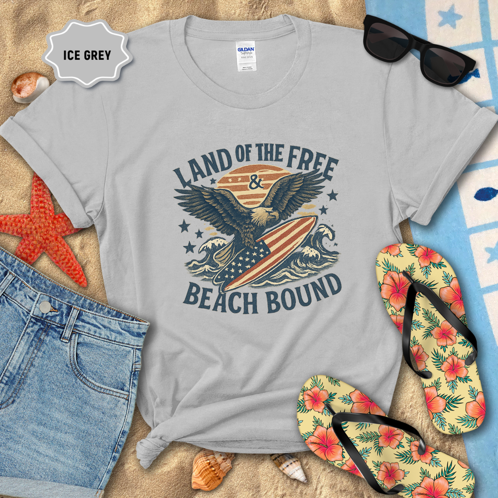 Gray t-shirt with eagle and surfboard design, sunglasses, flip-flops, and beach items on sand.