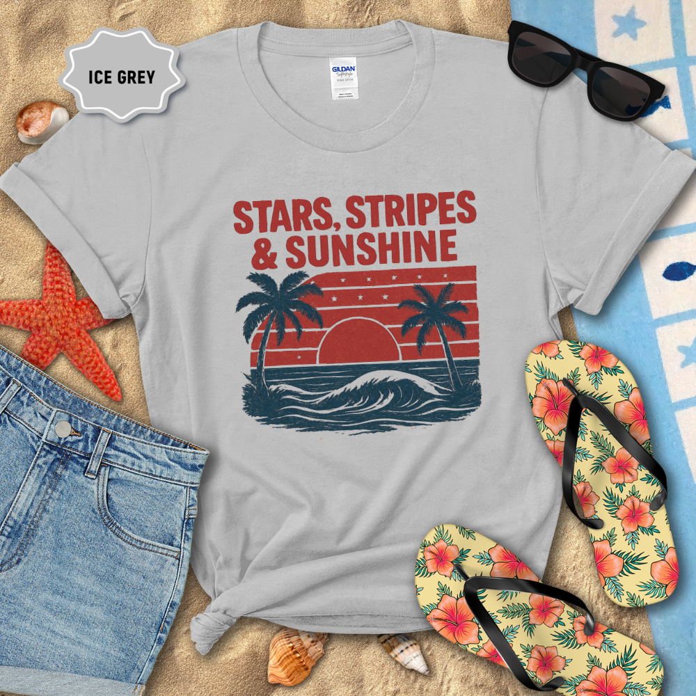 Gray t-shirt with 'Stars, Stripes & Sunshine' design on sand with beach accessories.