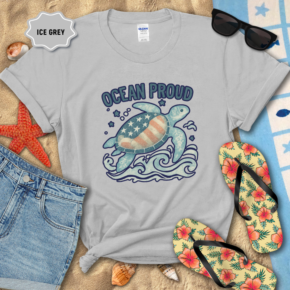 Gray t-shirt with turtle graphic and 'Ocean Proud' text on a beach setting with sunglasses, flip-flops, and denim shorts.