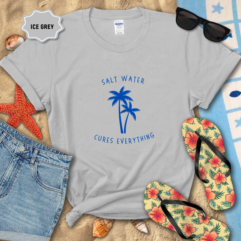 Gray t-shirt with 'Salt Water Cures Everything' design on sand with beach accessories.