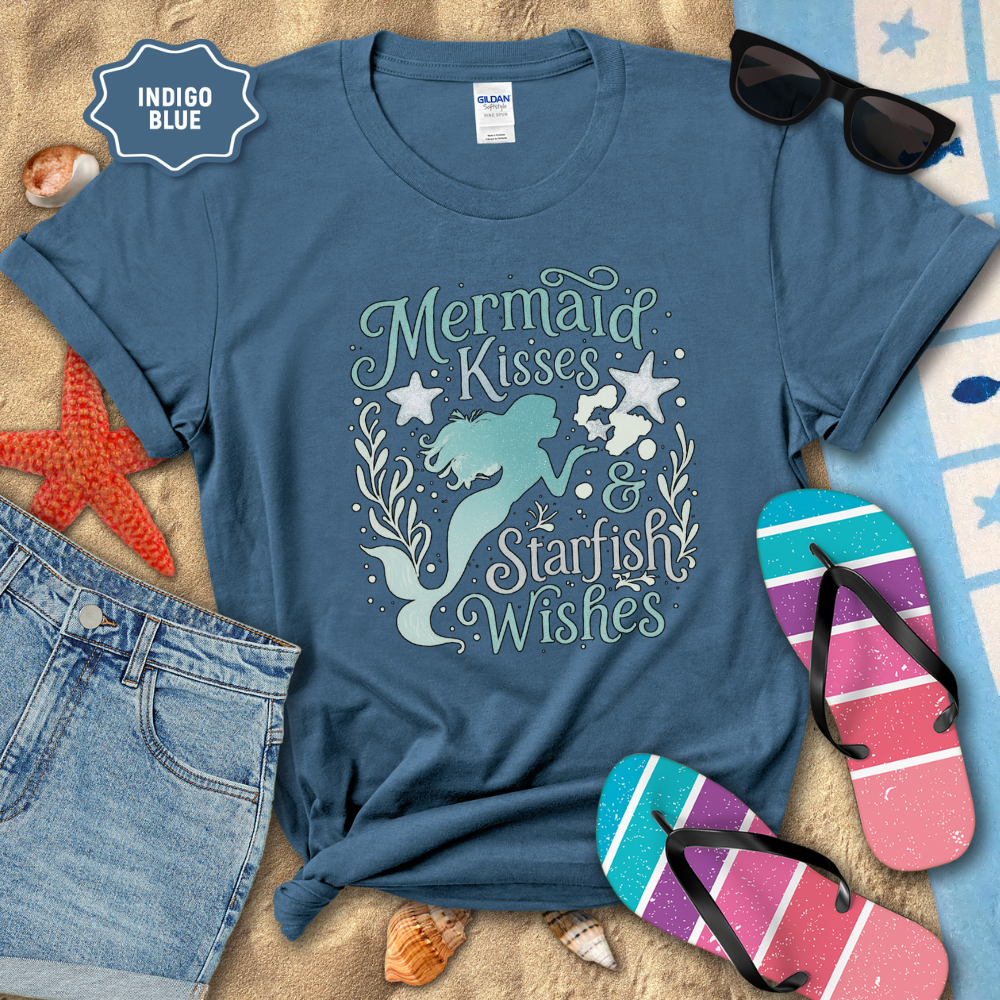Blue t-shirt with mermaid design and text, surrounded by beach accessories on sand.