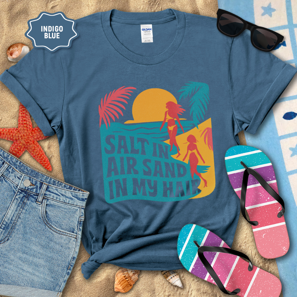 Indigo blue t-shirt with beach graphic and text, sunglasses, flip-flops, and denim shorts on a sandy background.