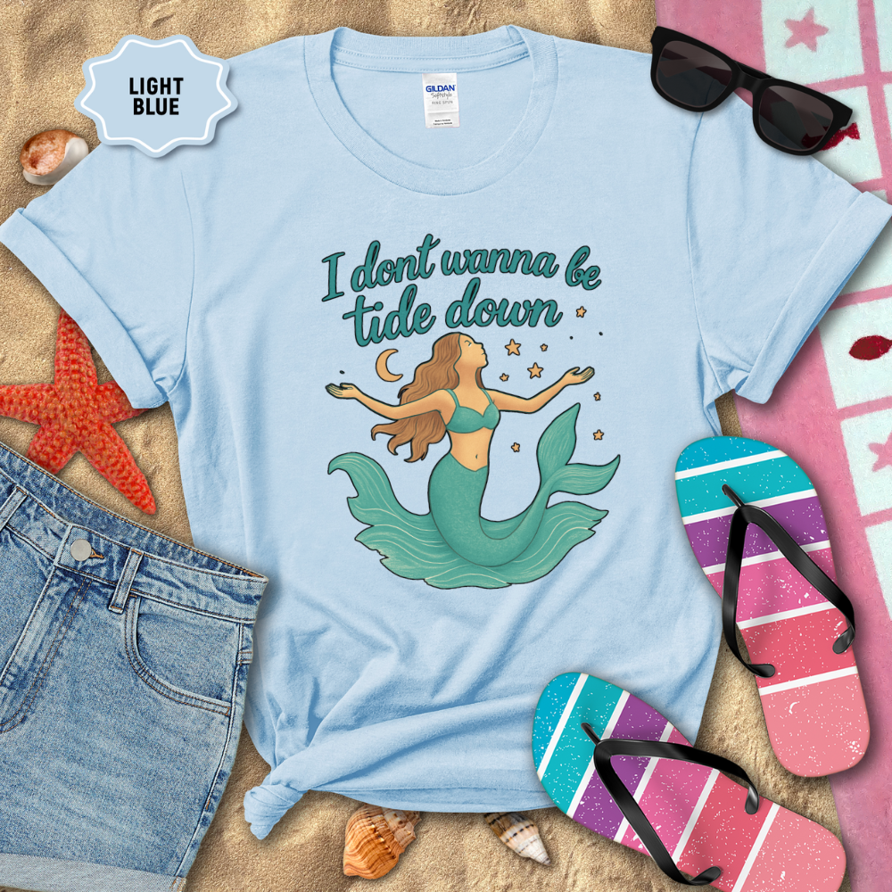 Light blue t-shirt with mermaid graphic and text on a sandy background with beach accessories.
